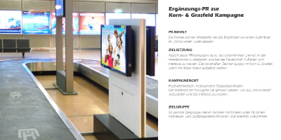 Werbescreen über einem Gepäckband, der das Minos‑Hotel und eine Familie zeigt.