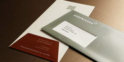 Briefumschläge der Agentur Augenzeuge: Vorderseite in Anthrazit mit weißem Logo, Rückseite in Cremeweiß mit QR-Code und eine braune Verschlussklappe an der kurzen Seite mit Adressdaten.
