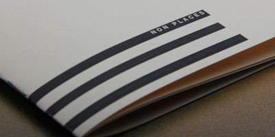 Nahaufnahme eines minimalistischen Buchcovers. Auf weißem, ungestrichenem Papier steht in kleiner, serifenloser Schrift der Titel ‚NON PLACES‘ über drei dicken, horizontalen schwarzen Balken.
