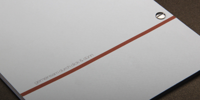 Rückseite des Magazins mit minimalistischer Grafik. Eine durchgehende orangefarbene Linie verläuft horizontal über die Fläche. Darüber platziert der kleingedruckte Satz „gemeinsam durch dick & dünn“. Die Bindung durch zwei silberne Buchschrauben ist deutlich erkennbar.