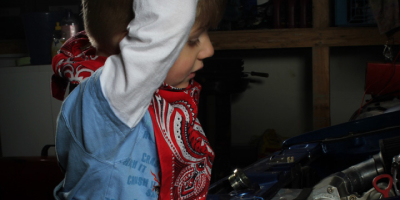 Nahaufnahme des Jungen von der Seite, wie er konzentriert unter die geöffnete Motorhaube eines blauen Autos blickt. Er stützt sich mit einer Hand am Rahmen ab. Im Hintergrund sind Werkstattregale mit Ölkanistern erkennbar.