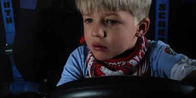 Close-up des Jungen, wie er hinter dem Lenkrad im Fahrersitz sitzt. Er hält das Lenkrad fest mit beiden Händen und blickt mit großen, leuchtenden Augen nach vorne, als würde er das Auto bereits über die Straße steuern. Die blauen Sicherheitsgurte rahmen ihn ein.