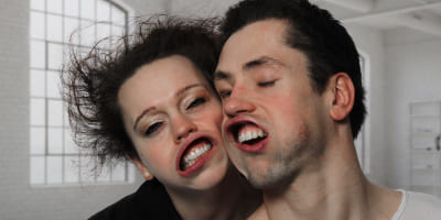 Zwei Personen in einem hellen Raum, deren Gesichter durch starke Luftströmung verzerrt sind, mit nach hinten gewehten Haaren.