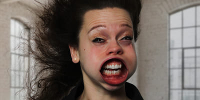 Eine Person vor großen Fenstern, deren Gesicht frontal durch starke Luftströmung verzerrt ist, mit nach hinten gewehten Haaren.