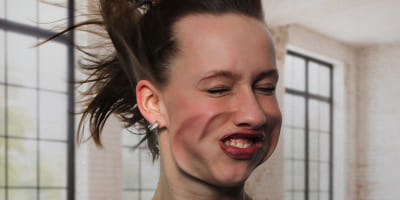 Eine Person in einem Raum mit großen Fenstern, deren Haare durch starke Luftströmung nach oben geweht werden, während das Gesicht deutlich verzerrt ist.