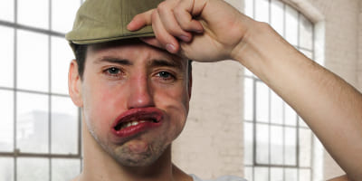 Eine Person mit Cap und T‑Shirt, deren Gesicht durch kräftige Luftkraft verzogen ist, während eine Hand den Schirm der Cap hält.