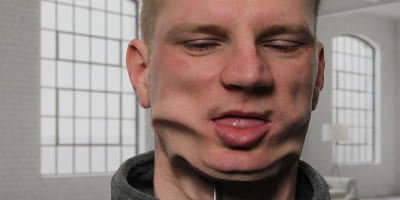 Eine Person in einem hellen Raum, deren Gesicht horizontal verzogen wirkt, mit herausgestreckter Zunge und verschobenen Gesichtszügen.