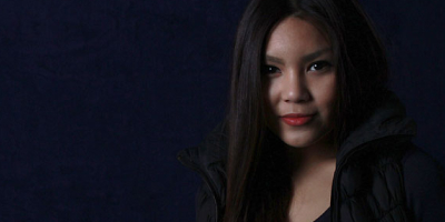 Eine Person mit langen dunklen Haaren steht vor einem dunklen Hintergrund, seitlich im Bild platziert. Das Gesicht und die Haare sind durch gerichtetes Licht hervorgehoben, während der Rest im Schatten liegt.