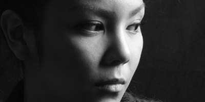 Ein Schwarz‑Weiß‑Portrait einer Person mit hochgesteckten Haaren, die seitlich aus dem Bild schaut. Starke Kontraste betonen Gesicht und Kleidung vor dunklem Hintergrund.