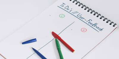 Handschriftliche Notizseite in einem Block mit der Überschrift „To Do’s // User-Feedback“ als Symbol für die Iterationsphase: Die Seite ist durch eine vertikale Linie in zwei Bereiche unterteilt, die mit einem grünen Smiley für positives Feedback und einem roten Smiley für Optimierungspunkte markiert sind. Drei farbige Stifte liegen bereit, um neue Arbeitspunkte für die nächste Prozessrunde zu dokumentieren.